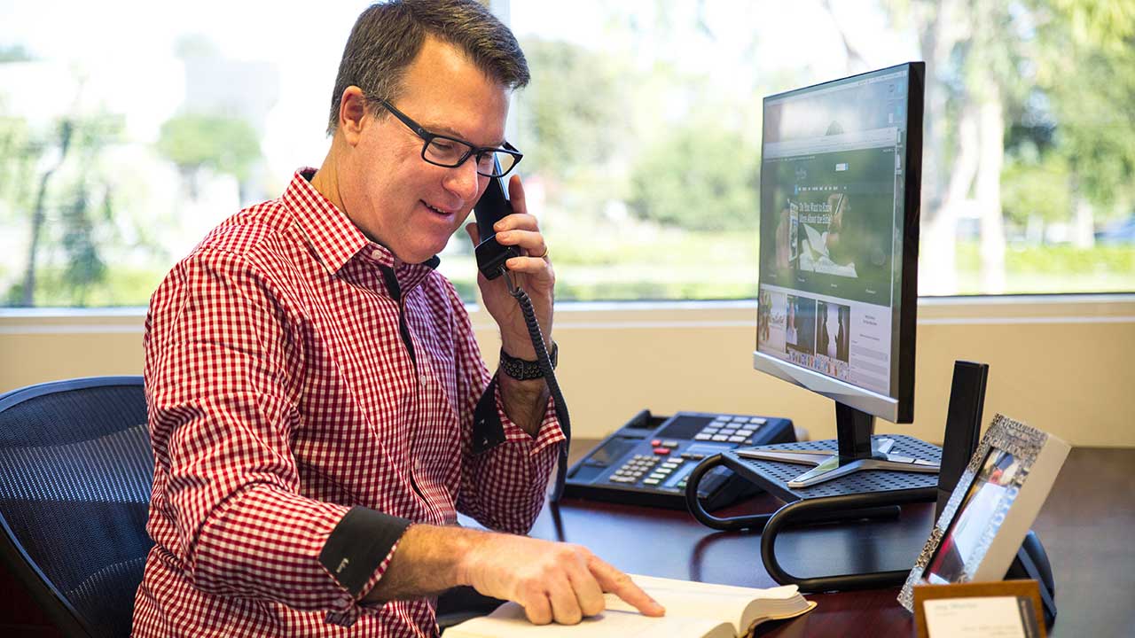 man talking on phone pointing to a page in his opened Bible on his desk